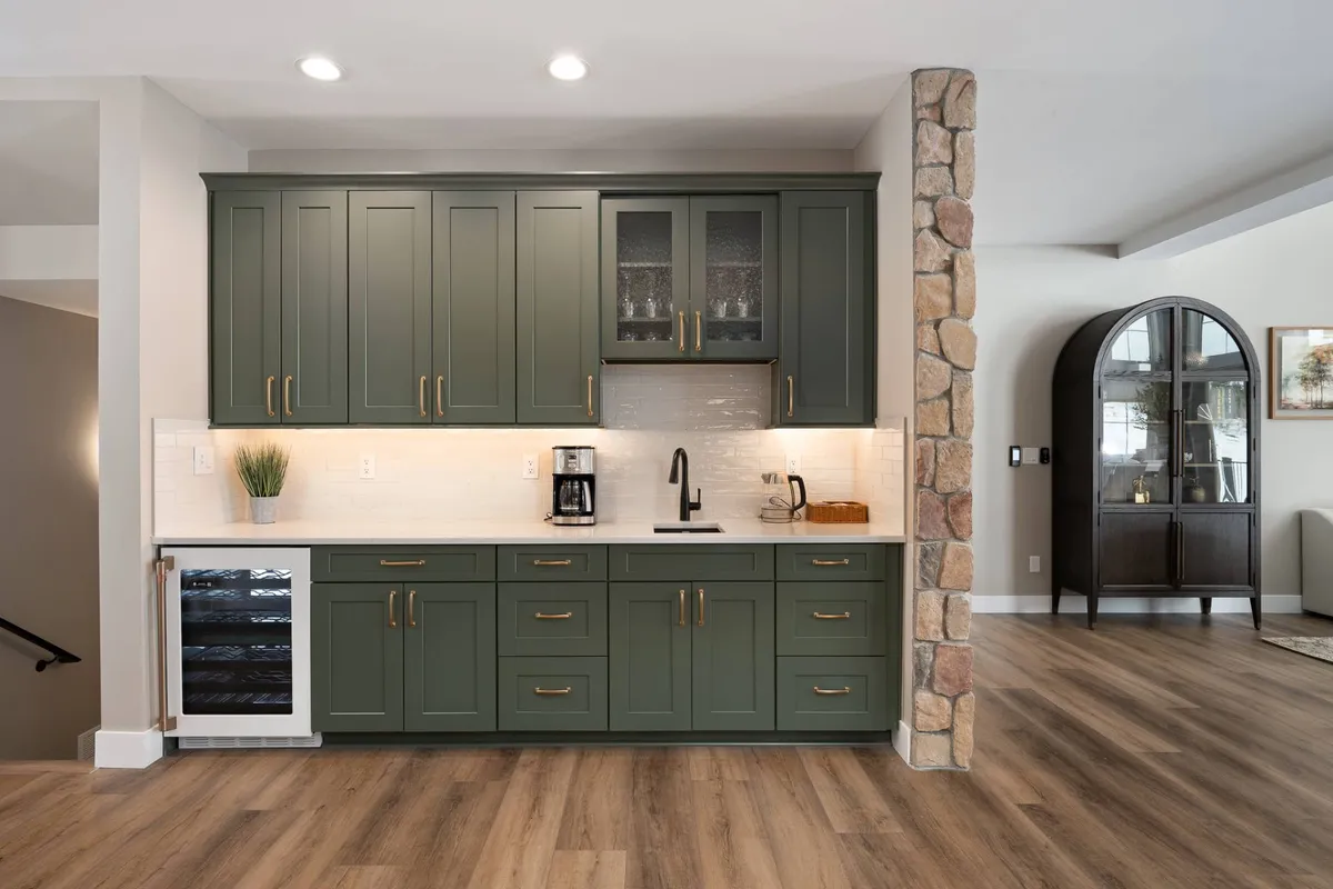 A stylish kitchen vignette emphasizing green cabinets, a stone column, and a countertop with a coffee maker and wine cooler—suggesting specialty items and the deluxe wet bar. The scene is set for both casual coffee breaks and evening celebrations.