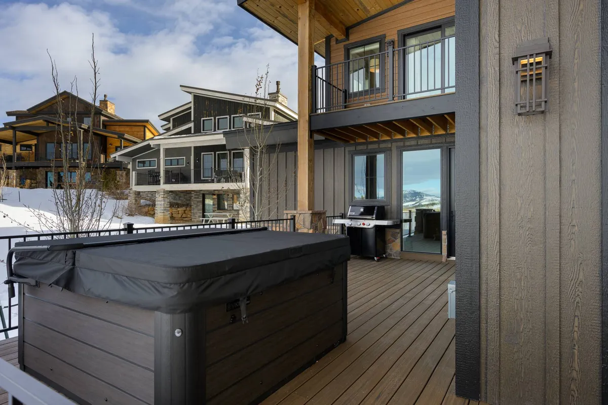 An expansive deck with a hot tub and wooden beams overlooks a tranquil landscape and distant mountains. The space is designed for gathering outdoors, with ample room for socializing or relaxing while enjoying sunset views.