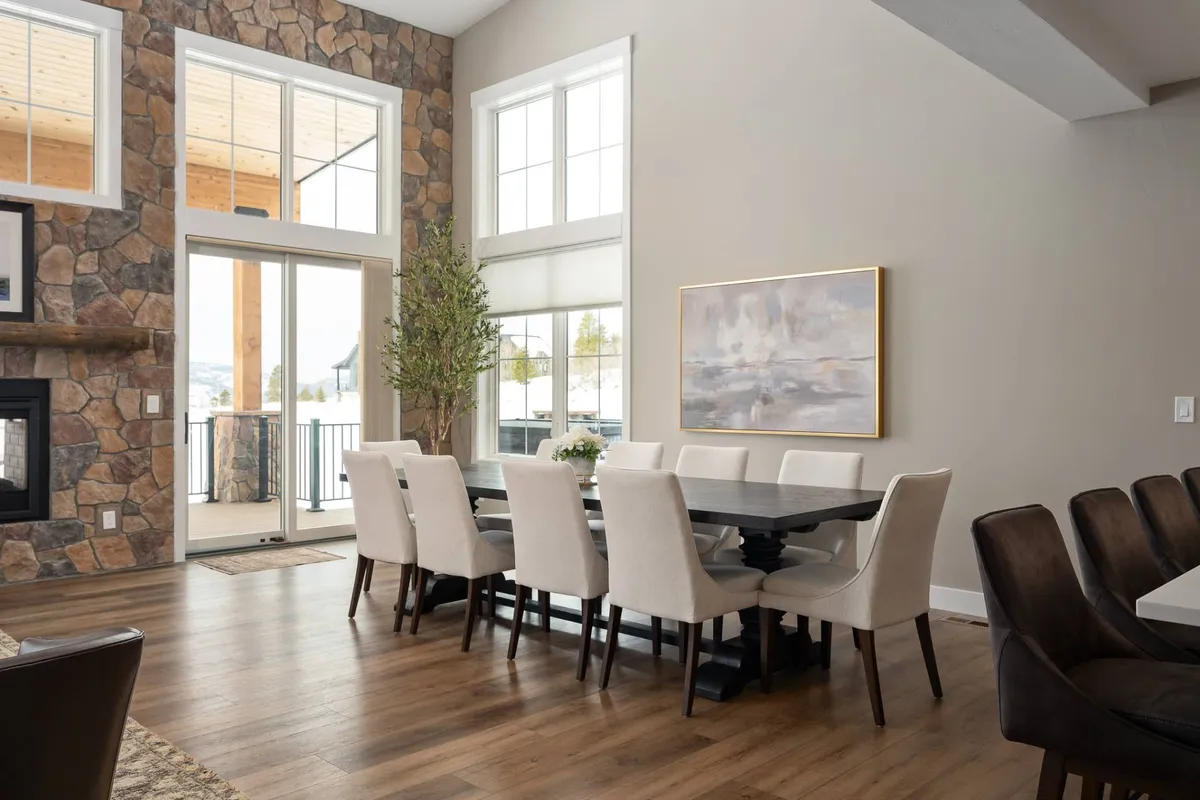 An inviting dining area anchored by a large wooden table, set before a rustic stone fireplace and expansive windows. The space is designed for family dinners, celebrations, and socializing, with natural light enhancing the warm, welcoming atmosphere.