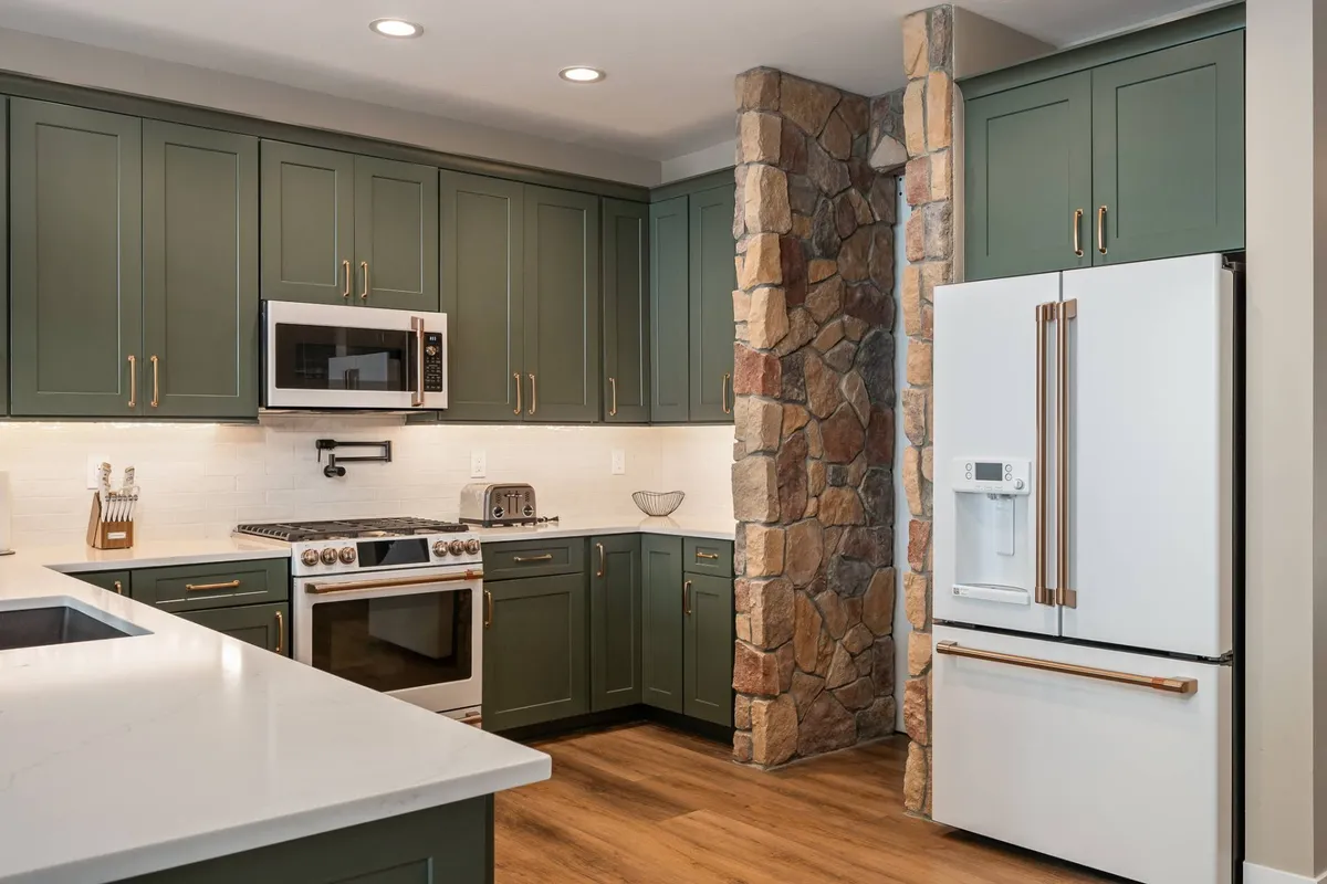 A spacious open kitchen with green cabinetry, a white stone countertop, a stone accent wall, modern appliances, and wood flooring. The setup encourages both cooking and entertaining, highlighting areas well-suited for prepping snacks or hosting guests.