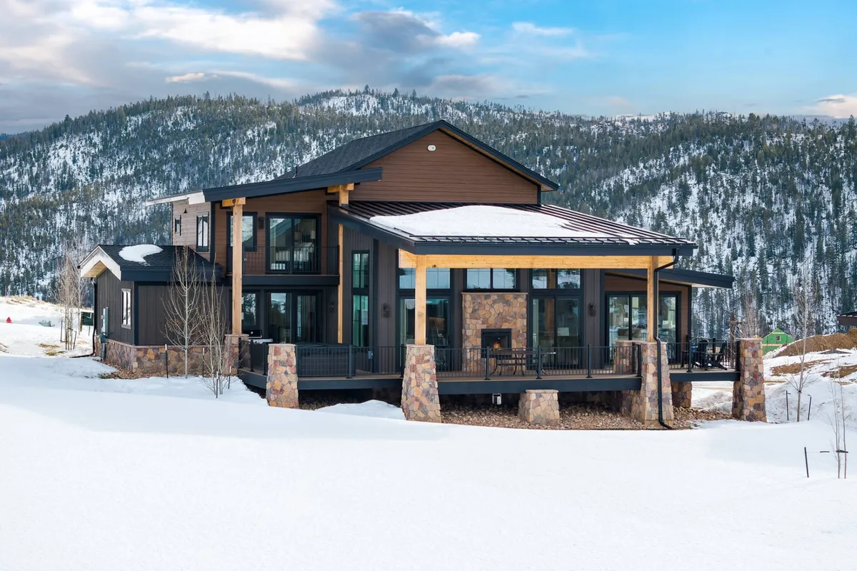The image shows a stunning, modern single-family home set against a snowy Granby, Colorado landscape. Large windows and stone pillars highlight the home's contemporary architecture, while the snow-covered surroundings evoke a sense of winter magic. The inviting exterior suggests warmth and space for family gatherings, with opportunities for skiing, snowboarding, and outdoor relaxation just steps away.