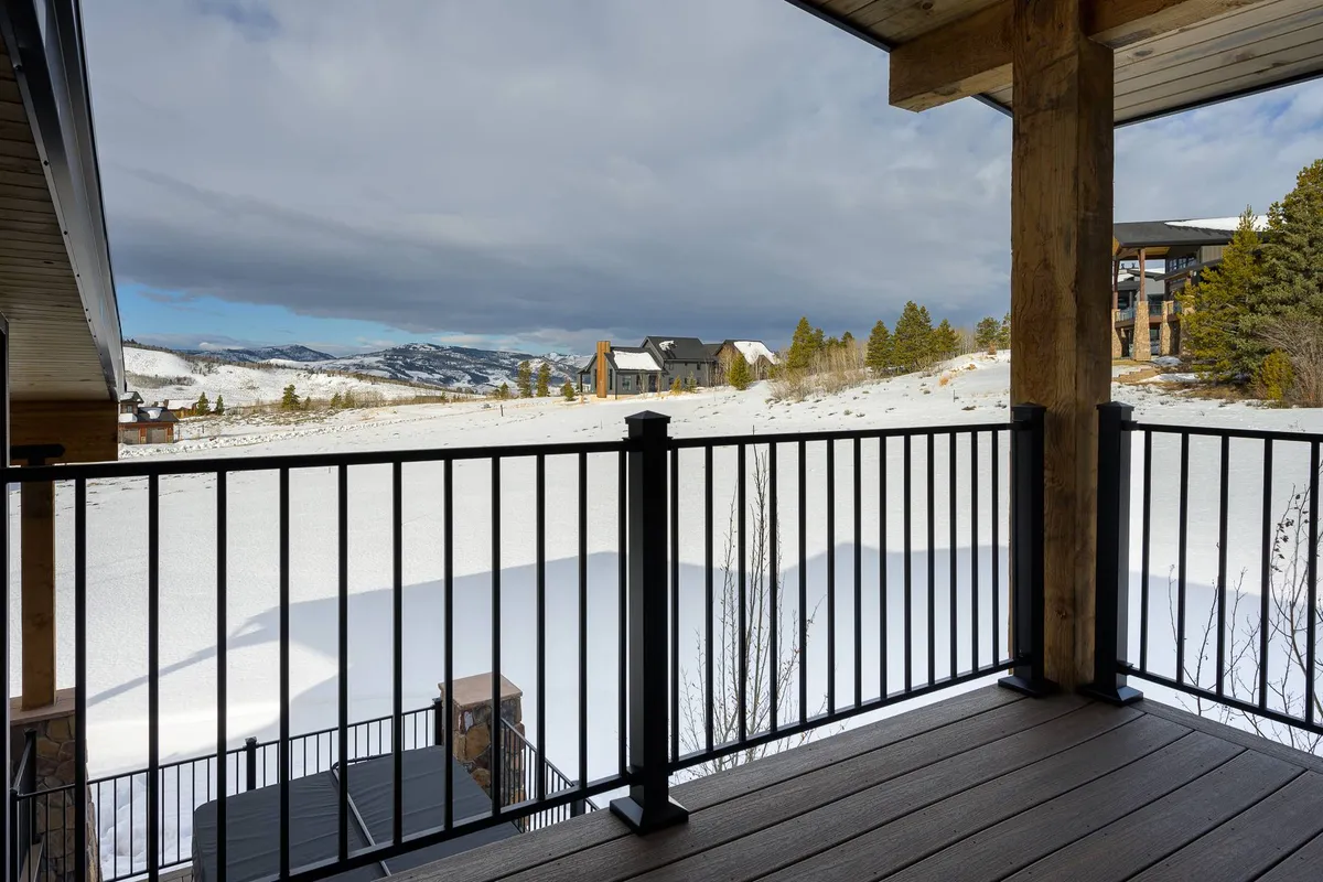 A tranquil view from a balcony or deck, looking out over a snow-covered landscape and distant mountains. The wooden railing and neighboring homes add to the sense of a welcoming resort community, perfectly situated for outdoor adventures.
