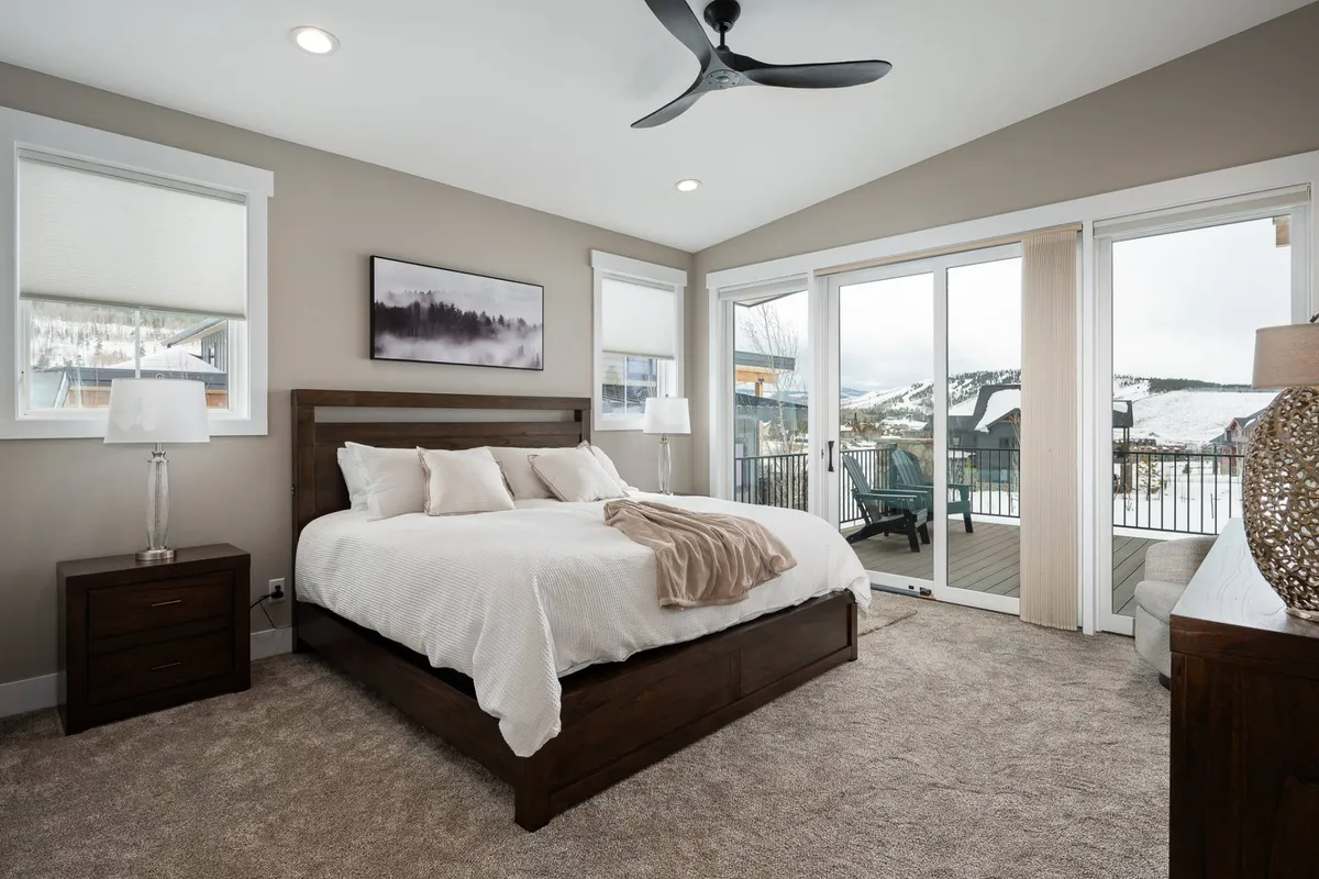 A serene bedroom featuring a bed with crisp white bedding, a nightstand with a lamp, and sliding glass doors opening to stunning mountain views. Soft natural light fills the room, emphasizing its calm and inviting atmosphere.