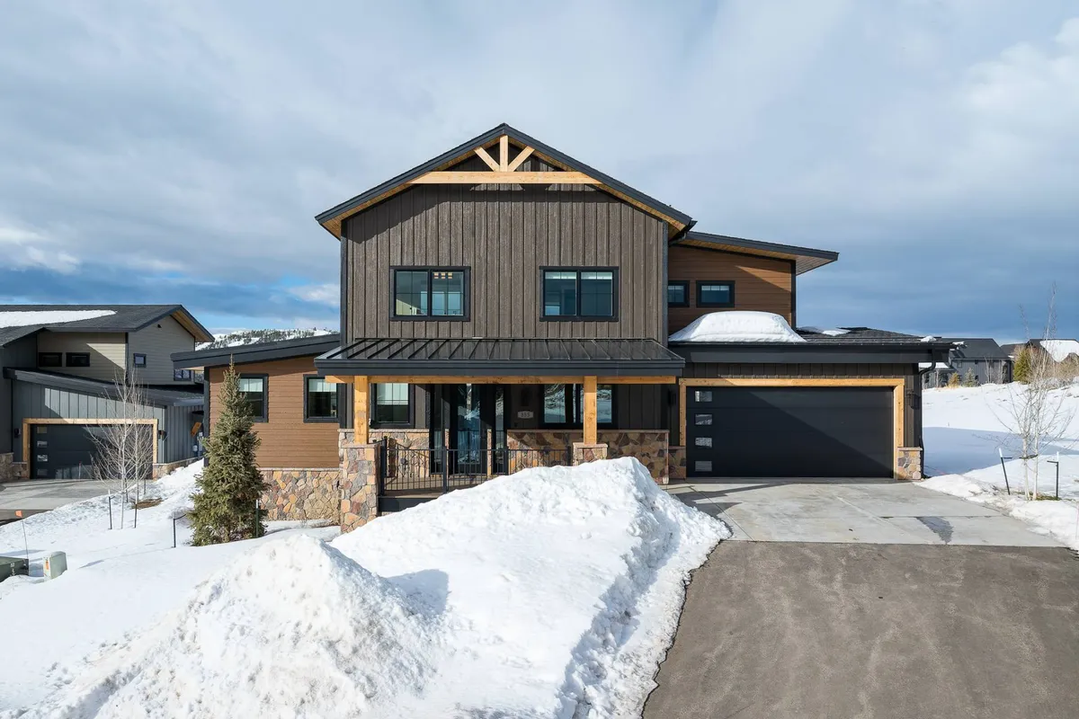 A modern home exterior featuring stone accents, large windows, and a snow-covered landscape. The inviting facade is complemented by a spacious porch, perfect for enjoying the mountain air. The scene evokes the warmth and coziness of mountain living, especially in the winter season.