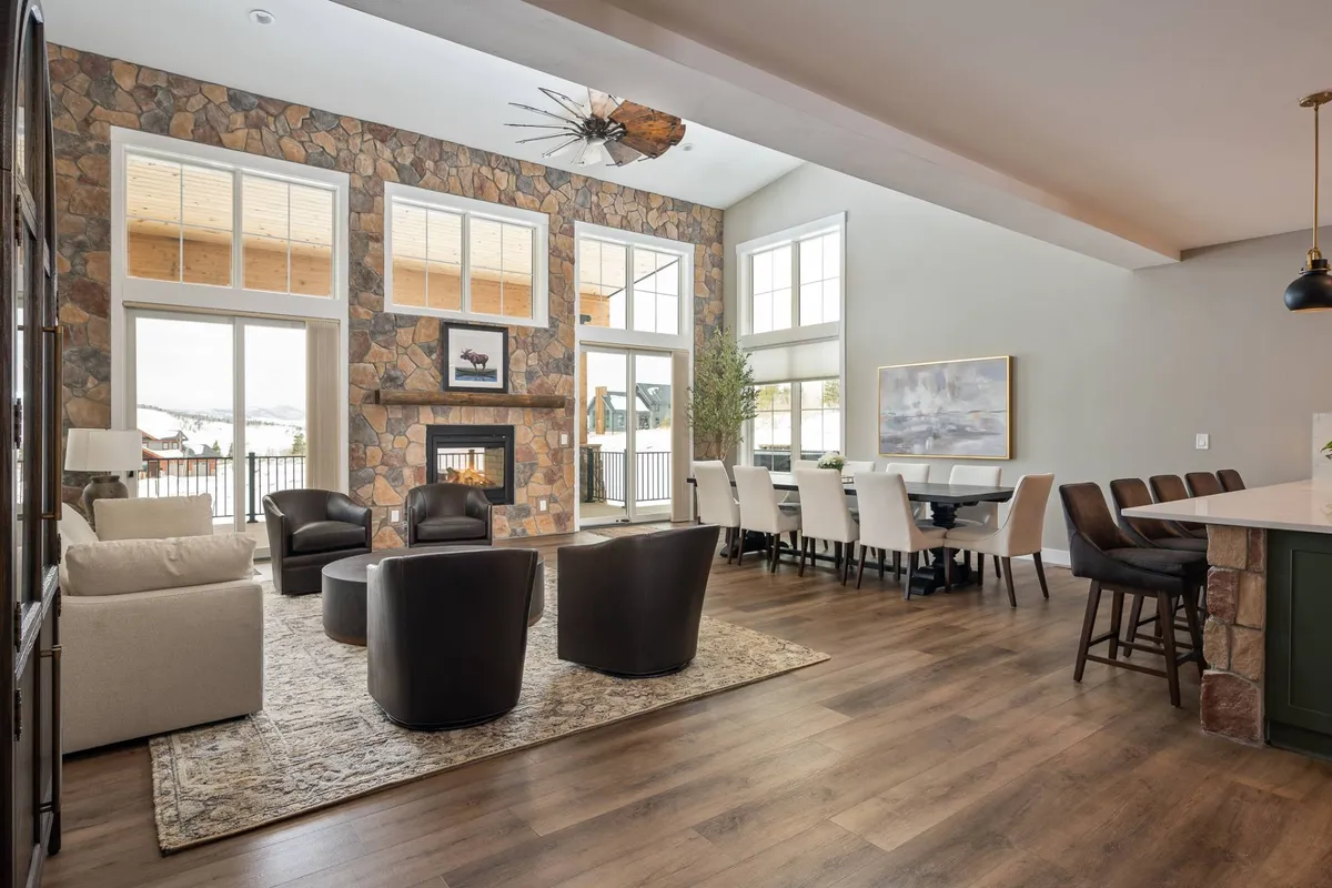 This inviting living space features a grand stone fireplace as its centerpiece, with comfortable seating arranged around it and expansive windows framing scenic mountain views. A dining table sits nearby, allowing for seamless transitions between dining and relaxing. Designer lighting adds a warm, welcoming glow throughout the space.