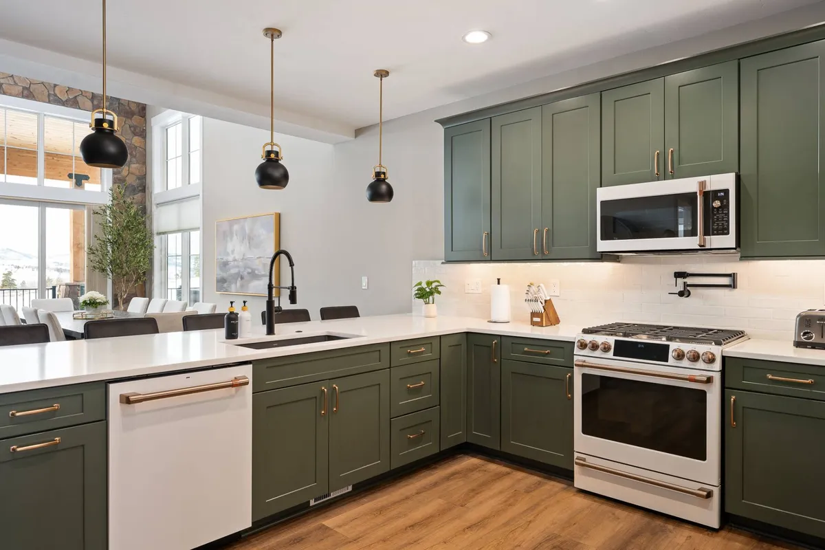 A bright, elegantly designed kitchen featuring sage green cabinetry, white quartz countertops, brass hardware, and black pendant lights. The kitchen is styled for both culinary activity and social gatherings, with plenty of workspace and a warm, welcoming atmosphere.